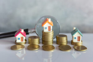 Small house resting on a pile of coins beside a magnifying glass.